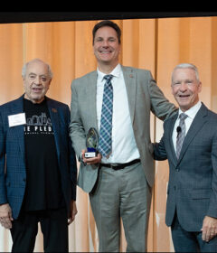 Three men stand on a stage and one holds an award.
