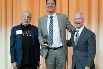 Three men stand on a stage and one holds an award.