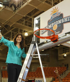 A woman holds a piece of net while standing on ladder underneath a basketball hoop.