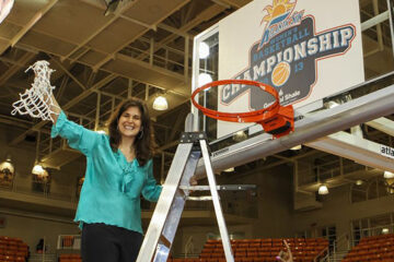 A woman holds a piece of net while standing on ladder underneath a basketball hoop.