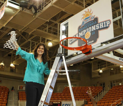 A woman holds a piece of net while standing on ladder underneath a basketball hoop.