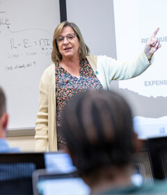 A female professor talks at the front of a classroom