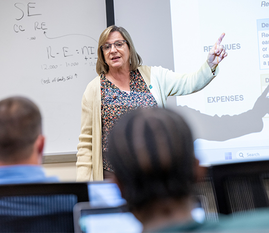 A female professor talks at the front of a classroom