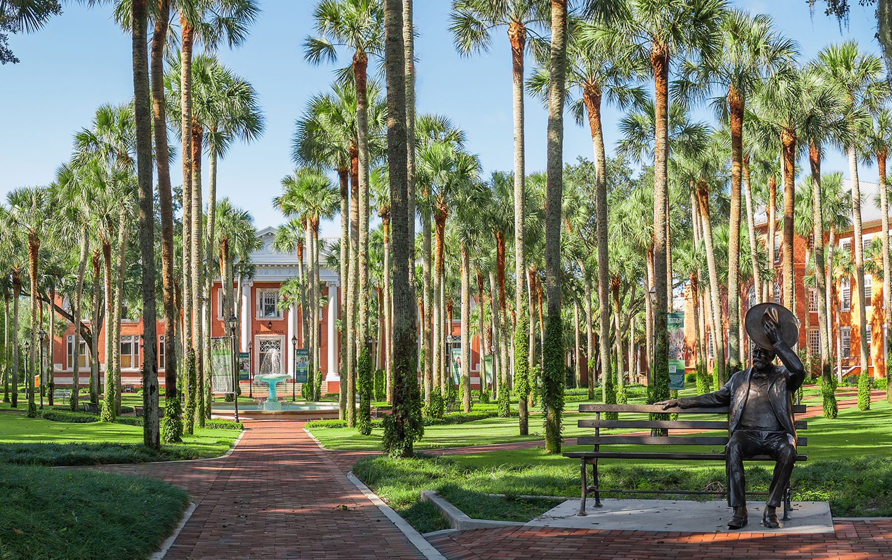 A seated statue of a man tipping a hat sits at the front of a pretty campus scene